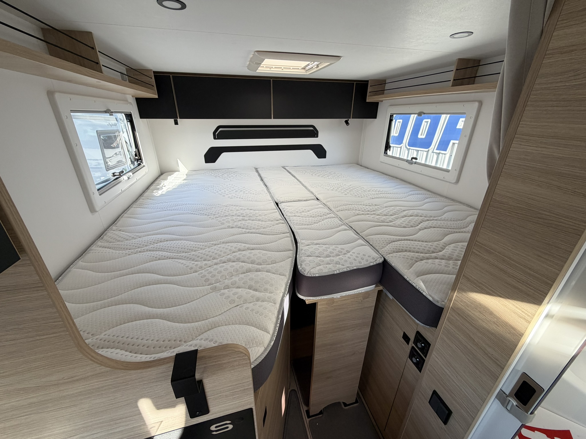 Interior of a modern caravan bedroom featuring two single beds with white mattresses, separated by a small gap, light wood panelling, overhead storage cupboards, and windows on both sides letting in natural light.